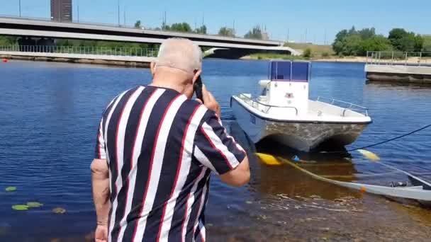 Gepensioneerde man met telefoon kijkt naar motorboot gelanceerd op river — Stockvideo