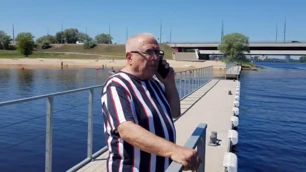 Un homme d'âge mûr discute avec un cellulaire sur une nouvelle jetée à la rivière bleue — Video