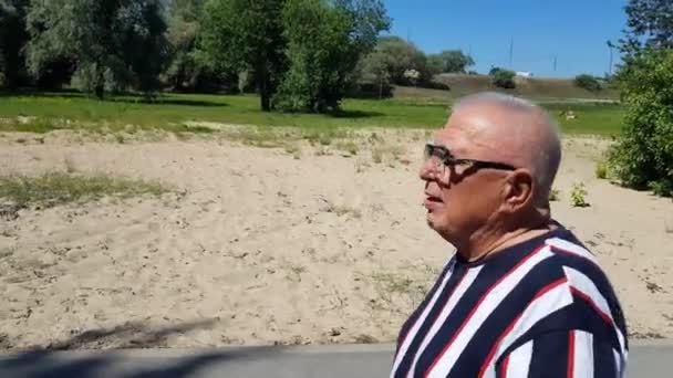 Hombre maduro en paseos en camiseta hablando a lo largo de la playa pasado parque. — Vídeo de stock