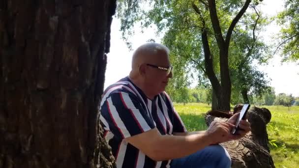 Érett férfi szövegek telefonon ül fa törzs a kertben — Stock videók