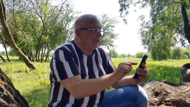 Ανώτερος άνθρωπος χρησιμοποιεί το τηλέφωνο κάθεται σε ένα τεράστιο πεσμένο δέντρο στο πάρκο — Αρχείο Βίντεο