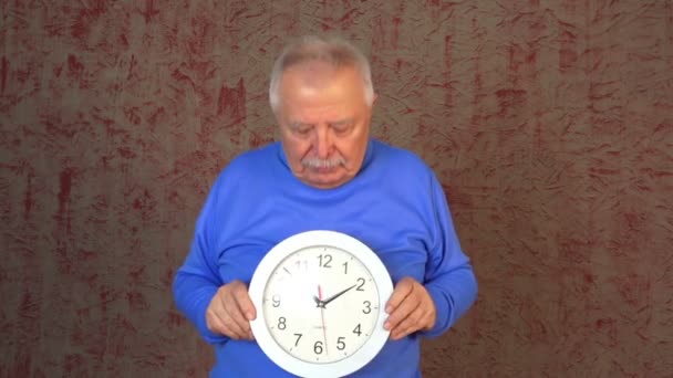 Пожилой человек в синей толстовке держит и обижается на часы — стоковое видео
