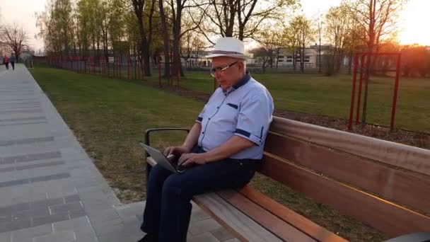 Pensionär i skjorta och jeans typer på laptop sitter i parken — Stockvideo