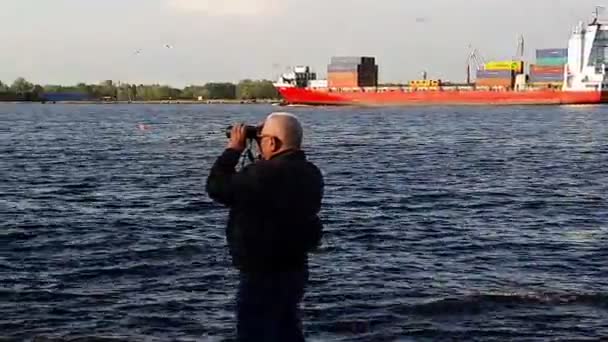 Man with binoculars on river bank against sailing cargo ship — Stock Video
