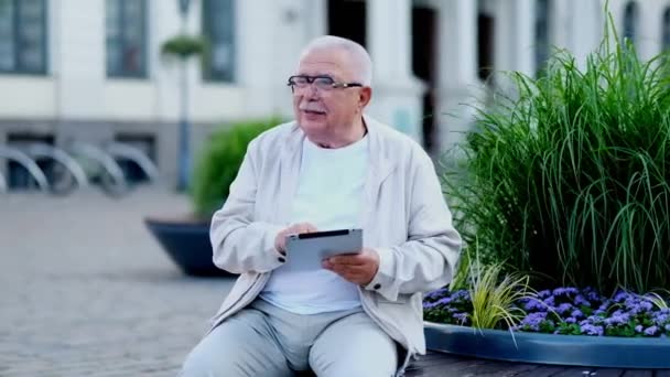 Hombre mayor con tableta se sienta en el macizo de flores en la plaza de la ciudad — Vídeos de Stock