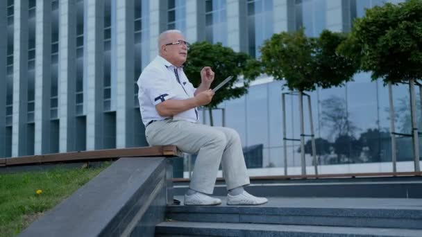 Jubilado alegre sostiene tableta sentado en banco de la calle — Vídeos de Stock