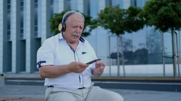 Pensionär med hörlurar lyssnar på musik på telefon på bänken — Stockvideo
