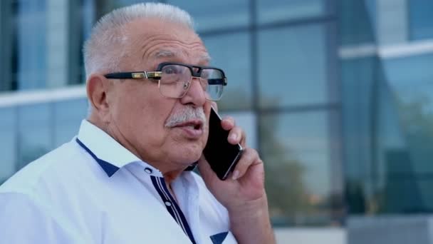 Pensionista habla por teléfono caminando a lo largo de edificio de oficinas — Vídeo de stock