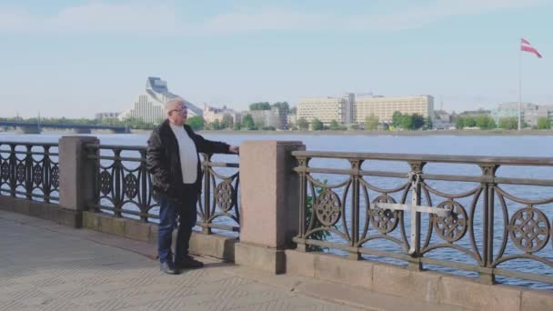 Penzista stojí na nábřeží řeky a má výhled na město — Stock video