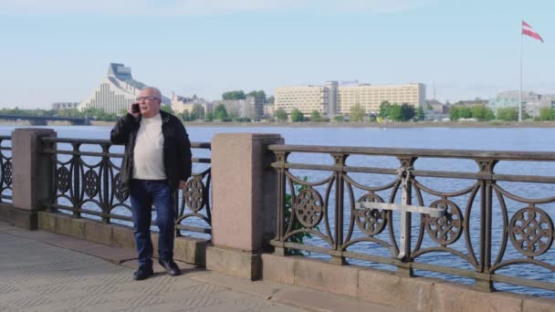 Пенсіонер в окулярах розмови по телефону стоять проти річки — стокове відео