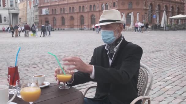 Старший человек в маске пьет коктейль за столом в уличном кафе — стоковое видео