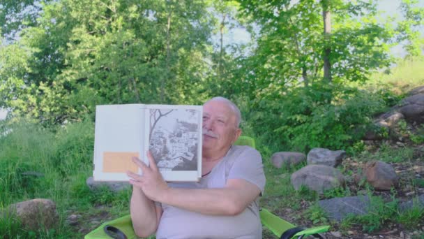 Feliz jubilado sonriente tiene libro de arte en las manos de cerca — Vídeo de stock