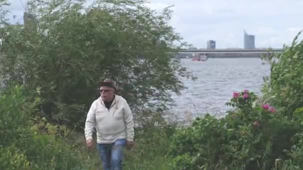 Hombre mayor con sombrero y gafas pasea por el parque cerca del río — Vídeos de Stock