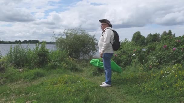Starší muž s batohem a taškou kráčí podél břehu řeky — Stock video