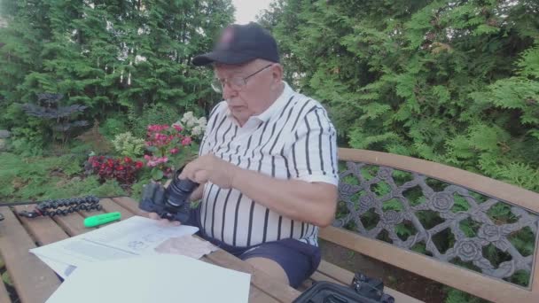 Senior man met bril reinigt cameralens aan tuintafel — Stockvideo