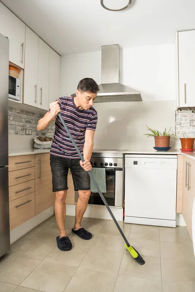 Man in casual clothes and slippers sweeping the kitchen floor. Full body.