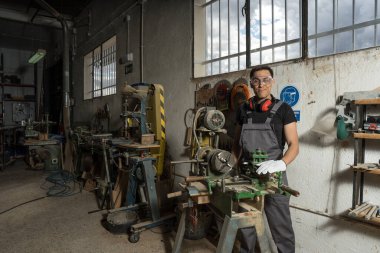 İşçi elinde koruyucu ekipmanla bir fabrikada gülümseyen kameraya bakıyor. Orta çekim.