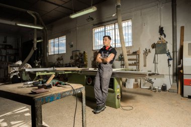 İşçi koruyucu aletlerle dikilip bir fabrikada gülümseyen kameraya bakıyor. Uzun atış, tüm vücut..