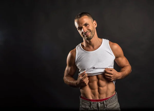 Athletic man lifting his shirt to show his abs. Mid shot. Black background.