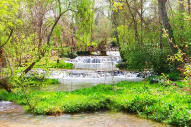 Turistler güzel bir bahar gününün tadını çıkarıyorlar. Manastırdaki yemyeşil bir parktaki şelalenin üzerindeki ahşap bir köprüde yürüyorlar.