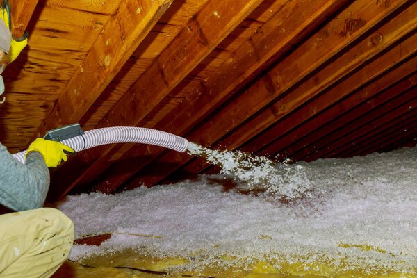 Technician spraying blown Fiberglass Insulation between Attic Trusses foam insulation repair tool in the white protect suit applies a construction foam from the gun to the roof.
