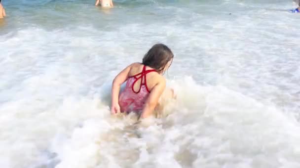 Bambina seduta sulla sabbia vicino all'oceano, le onde che schizzano nel suo corpo — Video Stock