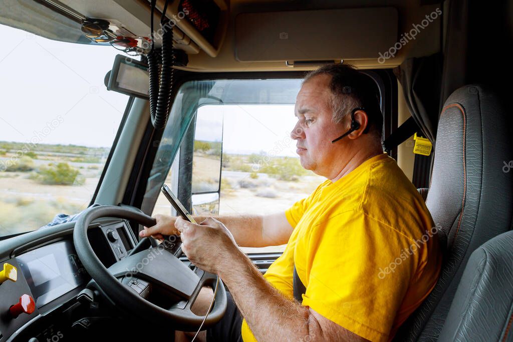 Driver in cabin on highway of smartphone in hand of man sitting behind the wheel of big modern truck vehicle