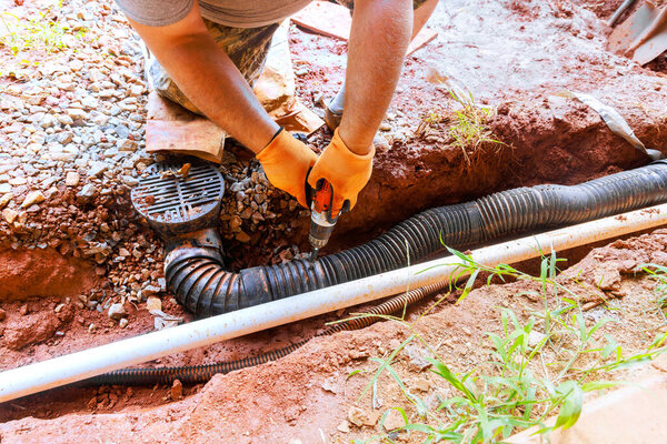 Contractor works on fixing drainage pipe in yard, wearing gloves using power drill tool on work day.
