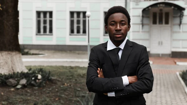 Young adult african american intelligent thoughtful business manager in suit and tie standing alone on the street near office and confidently looking at the camera. Image with copy space.
