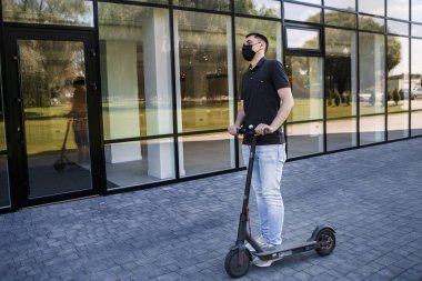 Elektrikli scooter 'da, scooter' da ya da e-scooter 'da müdürün tam boy portresi. Corona virüsünün ikinci dalgasından korunmak için maske takan bir adam ofis binasının yanında duruyor. Şehir ulaşımı.
