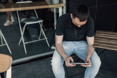 Kafenin dışında tablet bilgisayar ve kablosuz kulaklıklarla oturan genç serbest çalışanın portresini yakından çek. Adam salgını önlemek için koruyucu maske takıyor. Karantina ve korona virüsü kavramı 