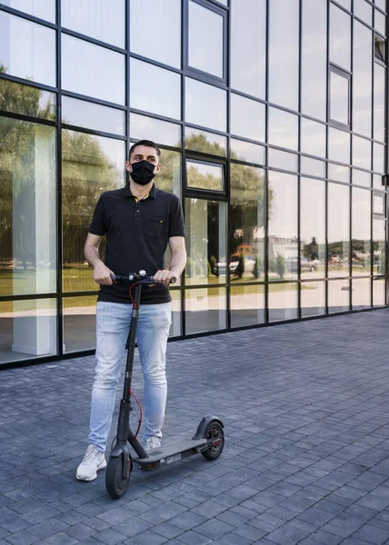Vertical full length portrait of student on electric scooter, kick scooter or e-scooter. Man wear mask to protect himself from second wave of epidemic corona virus. Urban transportation concept.