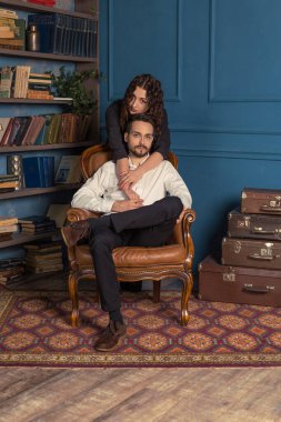 Eski antika odada zarif bir çiftin dikey portresi. Kadın ve erkek kitaplığın yanında poz veriyor. Eşiyle deri koltukta oturan modaya uygun sakallı zalim bir adam. Şehvetli, sevimli erkekler..