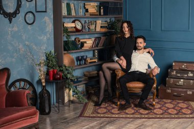 Eski antika odada genç ve zarif bir çiftin yatay portresi. Bir sürü antikayla kitaplığın yanında poz veren kadın ve erkek. Koyu kıvırcık kadın, kocasıyla birlikte koltukta oturuyor..