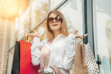 Çekici bir bayanın seçici odağı. Bir çok çanta alışverişi yapan kadın güneş gözlüğü takıyor. Beyaz gömlek giyen kız yeni kıyafetler taşıyor.