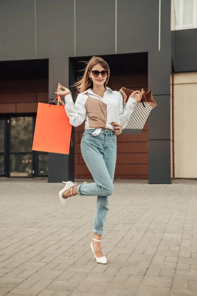 Avrupai bir kadının dikey portresi, güneş gözlüğü takıp alışveriş merkezinin yakınındaki beyaz gömlek kot pantolon mağazasından alışveriş yaptıktan sonra siyah cuma sezonunun arka planında, renkli çantalarla el ele tutuşan kadın.