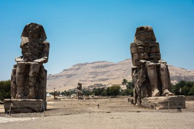 Luxor, Mısır - 27.07.2025. Memnon Colossi olarak bilinen iki devasa taş heykel Nil Nehri 'nin batı kıyısında arka planda kayalık bir dağ sırasına sahip uzun boylu dururlar.