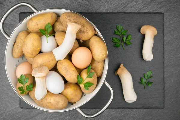 Mushroom Tortilla ingredients with raw potatoes, eggs, king oyster mushrooms and parsley leaves  in a white enamel sieve on a slate slap. Flat lay.