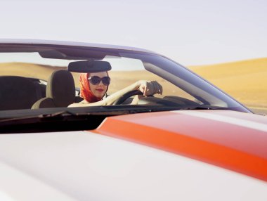 Sürücüdeki çekici kadın, spor, üstü açık, eski model bir arabanın direksiyonunda oturur. Güneş gözlüklü, başında kırmızı ipek bir eşarp, Dubai çölünde lüks bir yaşam tarzı.