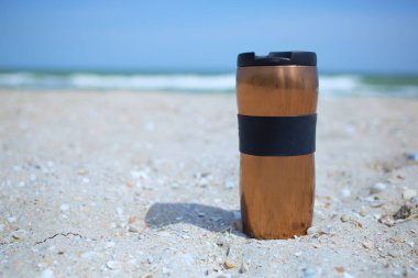 Bir bronz metal termos bardağı 300 ml kumda, deniz manzaralı sıcak ve soğuk kahve, çay içeceği, sahilde sabah içkisi, hiç kimse, sakin neşeli tatil konsepti. 