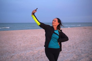 Çalışan giysiler içinde çekici bir kadın koşuyor, güzel deniz sahilinde koşuyor, fotoğraf çekmek için duruyor, video görüşmesi, aktif tatil konsepti.