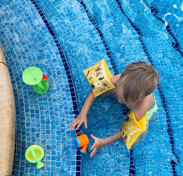 Little blond boy play with plastic colorful toys at swimming pool with blue small tile and stairs, wearing inflatable armbands, summer playful background, family vacation weekend, top view