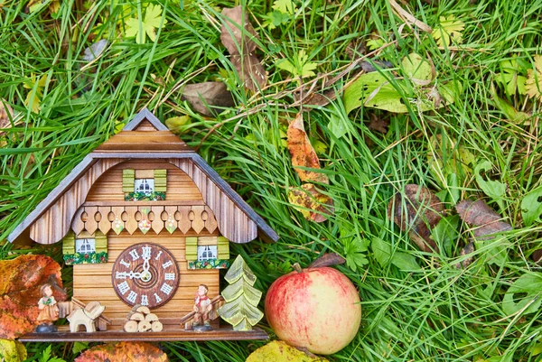 Eine hölzerne Wanduhr in Form eines Hauses, ein Apfel und gelbe Blätter liegen auf dem hellgrünen Gras. Blick von oben. — Stockfoto