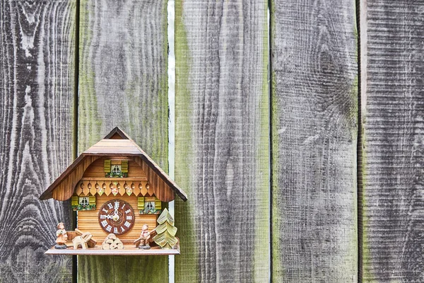 Eine hölzerne Wanduhr in Form eines Hauses hängt an alten grauen Brettern mit grünem Moos. Gestaltungselement. — Stockfoto