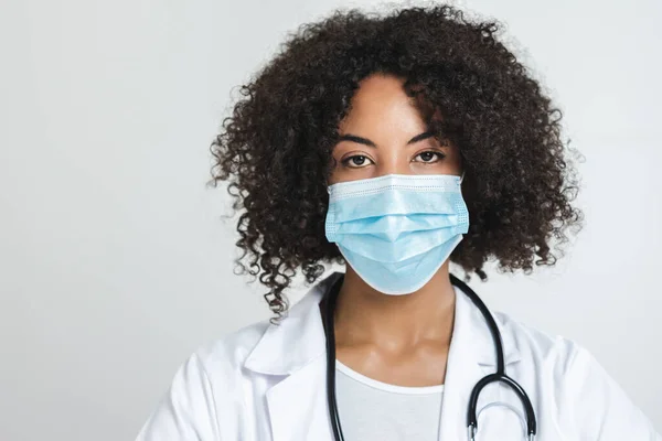 Beyaz arka planda cerrahi maskeli Afro bir kadın doktor portresi.