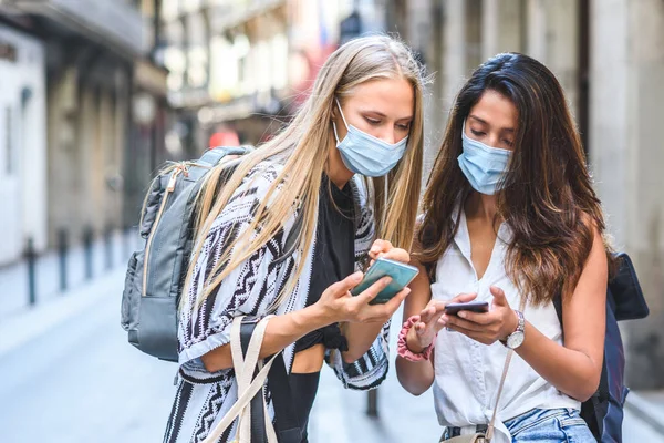 Maskeli ve sırt çantalı iki genç bayan turist şehrin sokaklarında cep telefonlarını kontrol ediyor.
