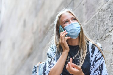 Güzel sarışın beyaz bir kadının portresi mavi bir maske takıyor ve eski bir binanın yakınında telefonla konuşuyor. Boşluğu Sola Kopyala