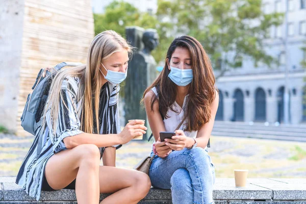İki güzel bayan arkadaş sokakta taş bir bankta oturuyor ve birbirlerine akıllı telefon ekranlarını gösteren mavi hijyenik maskeler takıyorlar.