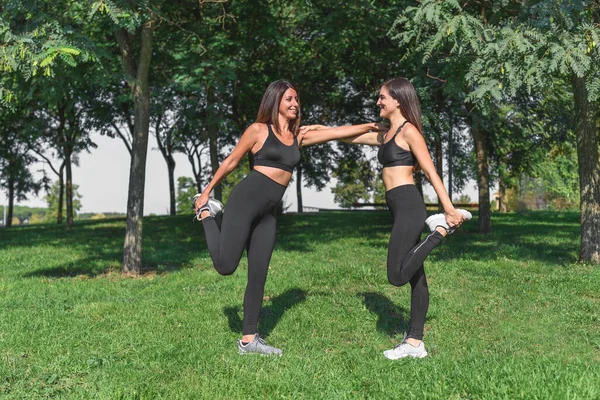 caucasian and latin female friends helping each other to keep equilibrium while stretching their legs near trees on a green park
