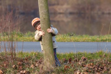 Küçük çocuk bir ağacın arkasından dışarı bakıyor.